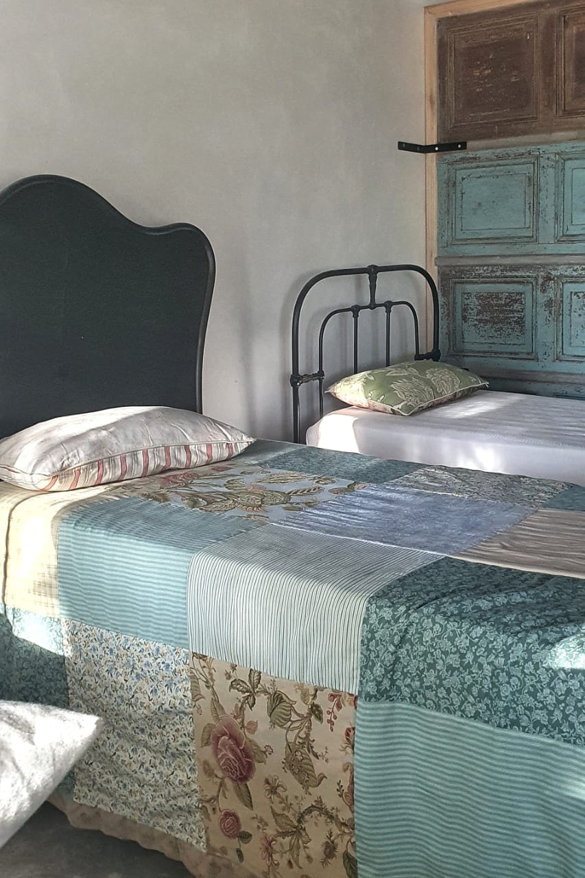 A cozy sleeping area features two beds with patchwork coverings in soft colors. Light filters in, illuminating the textures of the fabric. A vintage wooden door with an intricate design serves as a backdrop, adding character to the space.