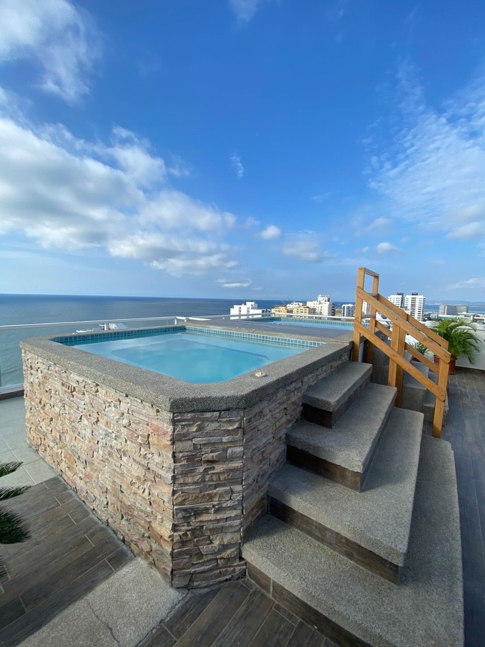 A stone-accented jacuzzi is positioned on an elevated terrace, offering panoramic views of the ocean and city skyline. Sturdy wooden steps lead up to the jacuzzi, where vibrant blue waters reflect the clear sky.