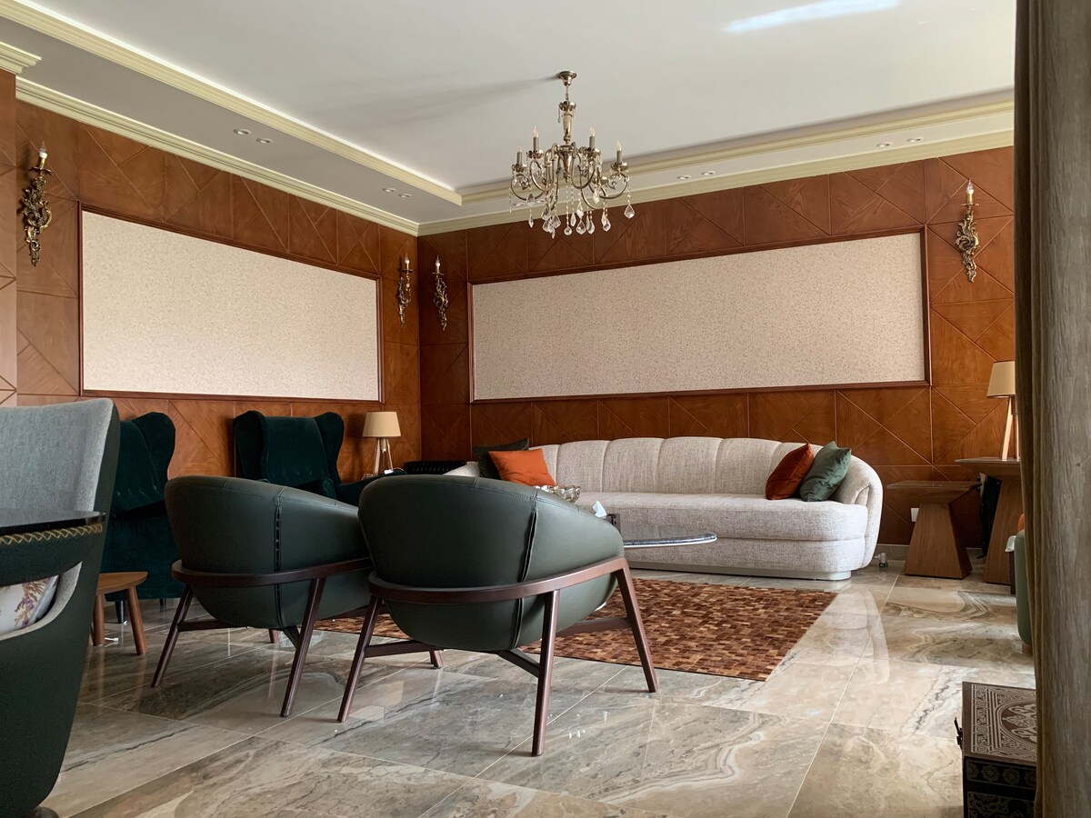 A stylish living area features a soft beige sofa paired with dark green armchairs. A decorative chandelier hangs from the ceiling, and a large area rug lies beneath a coffee table. The walls are accented with warm wood tones and textured panels.