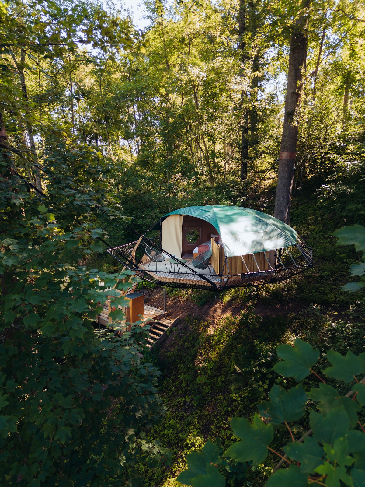 A circular accommodation rests on a raised platform amidst a wooded area. Featuring a green canopy, the structure is surrounded by lush foliage. Steps lead to the entrance, providing a serene connection to nature.