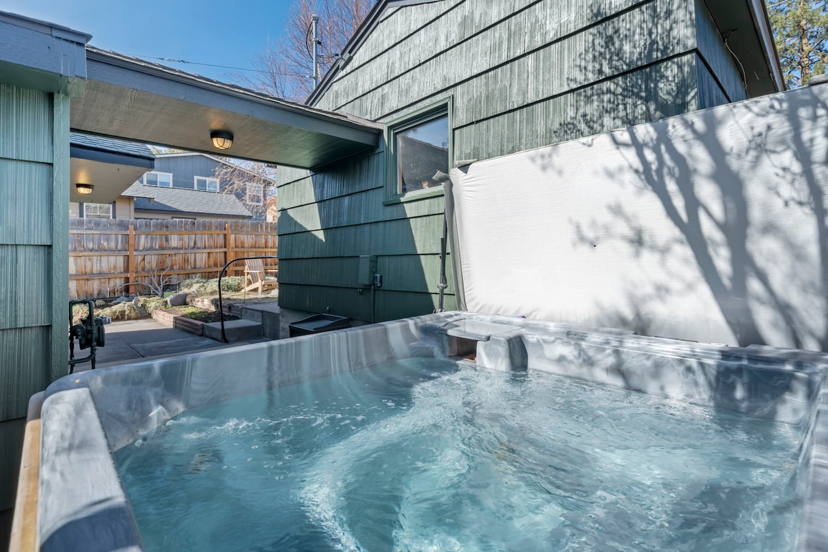A private salt-water hot tub is positioned outside, surrounded by green siding and a concrete patio. Sunlight reflects off the water's surface, creating a tranquil atmosphere. Landscaping elements are visible in the background, offering a serene environment for relaxation.