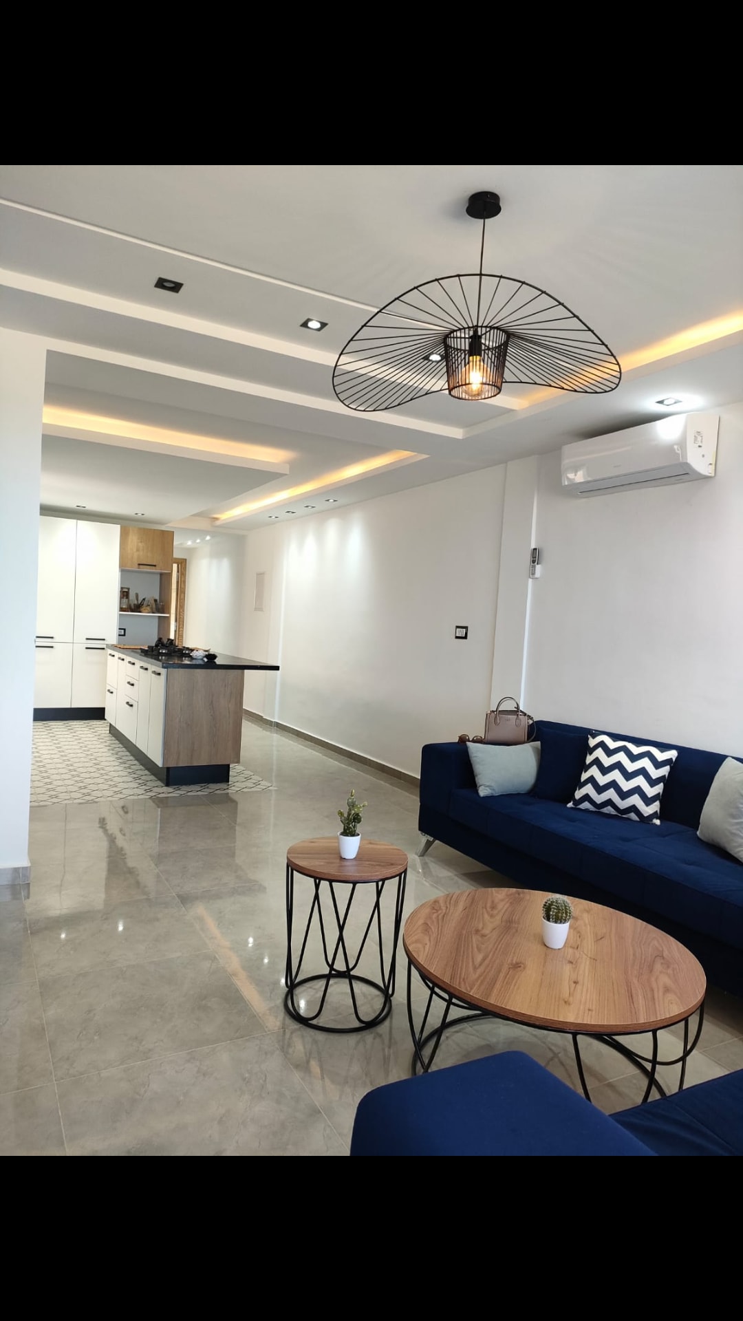 A contemporary living space features a blue sofa and two wooden coffee tables. An open kitchen with a central island is visible, complemented by warm lighting and a minimalist design. Air conditioning is installed on the wall, contributing to the comfortable atmosphere.