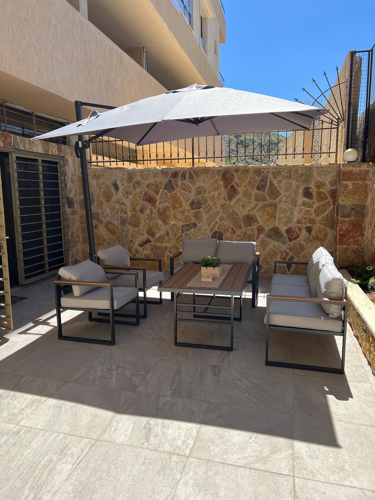 An outdoor seating area is presented, featuring a table surrounded by four light-colored armchairs. A large umbrella provides shade, and natural stone walls enhance the privacy. Sunlight illuminates the space, highlighting the clean tiled floor.