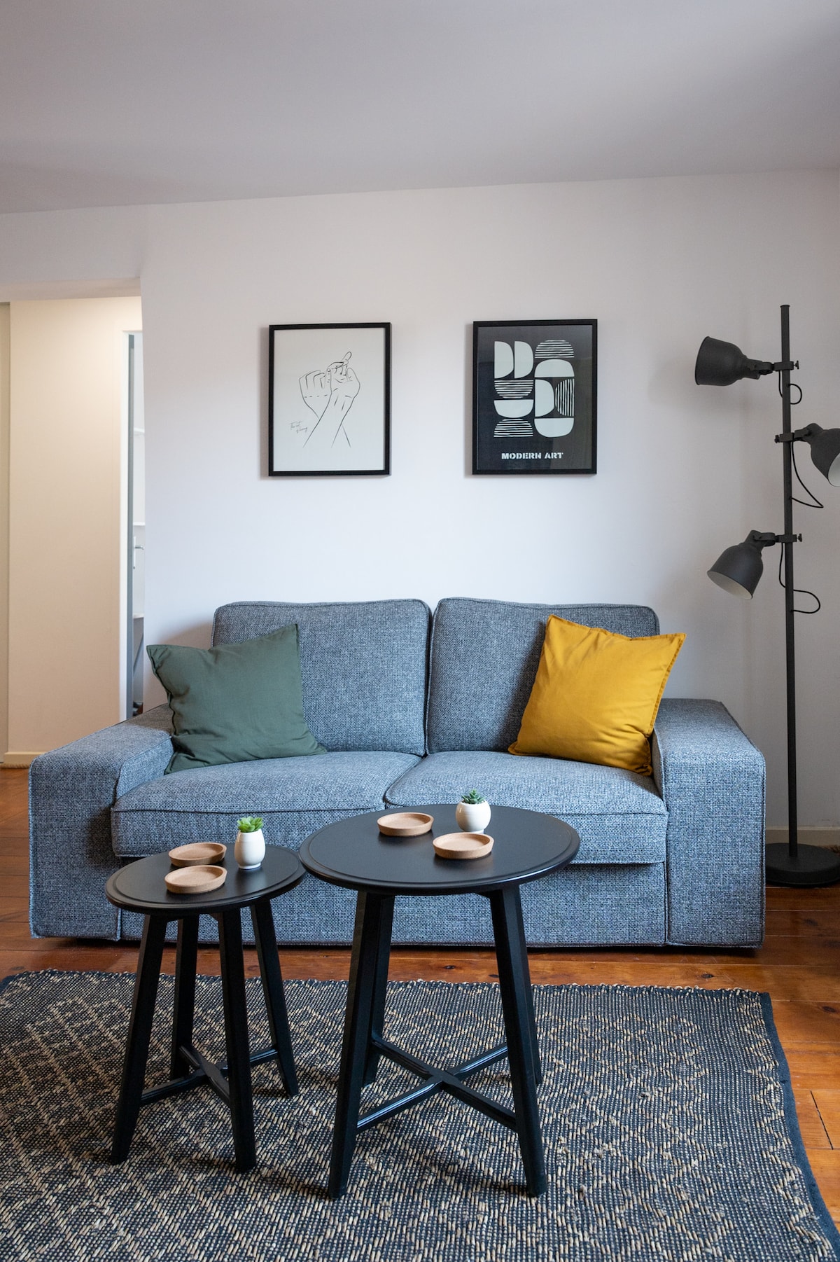 A cozy seating area is presented with a gray sofa adorned with two decorative pillows, one in green and one in yellow. Two small black tables are positioned in front, featuring minimalistic decor items. Art prints are showcased on the wall beside the sofa.
