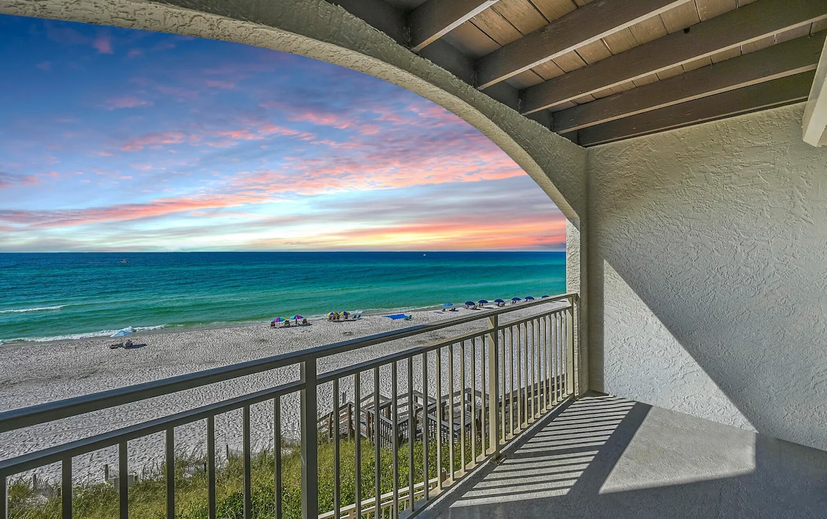 A balcony view captures the tranquil Gulf of Mexico, where soft waves kiss the sandy shore. A colorful sunset casts hues across the sky, enhancing the beach scene below. The railing frames the vista, inviting relaxation in this coastal setting.