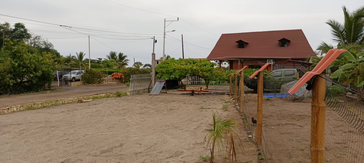 A sandy area is framed by wooden fencing, with sparse vegetation and a long structure visible in the background. A sloped ramp and space for activities are present, creating a versatile outdoor environment. Surrounding palm trees and tropical plants enhance the natural setting.