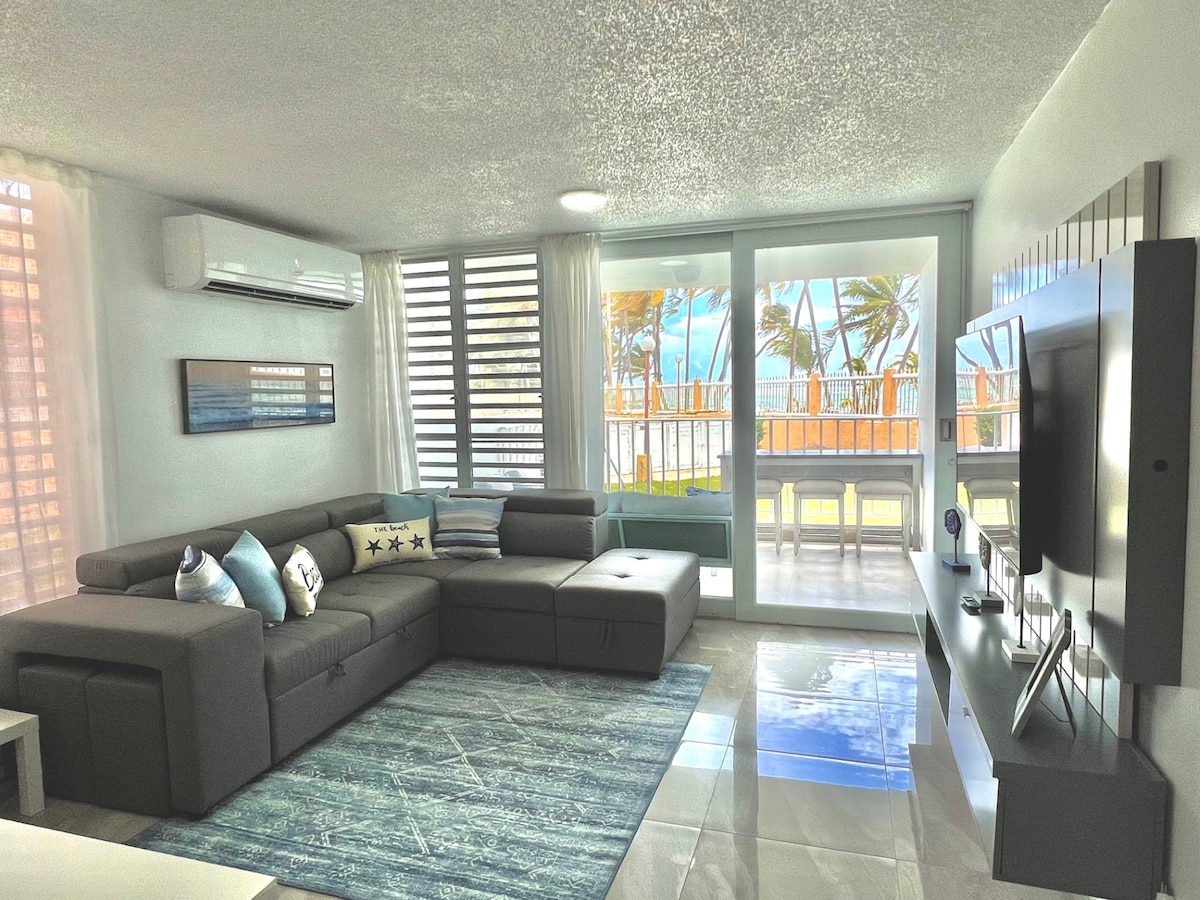 A modern living area features a large sectional sofa adorned with decorative pillows. Natural light filters through sheer window treatments, illuminating a soft area rug. Sliding glass doors offer a view of palm trees and the outdoors, enhancing the spacious feel.
