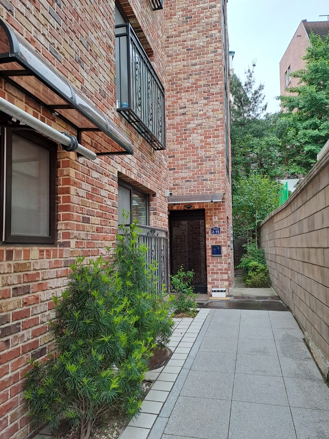 A narrow pathway leads to the entrance of the building, flanked by neatly trimmed greenery and brick walls. The textured brick exterior contrasts with the smooth stone pathway, creating a welcoming approach. Soft natural light filters through the surrounding trees.