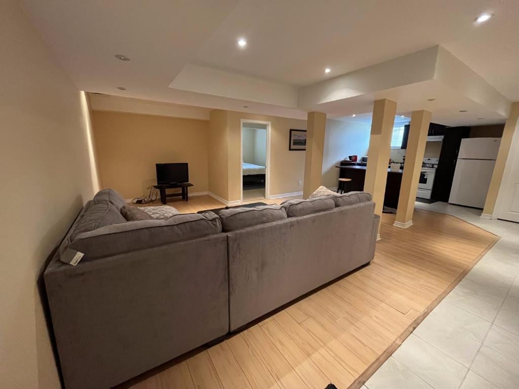 A spacious living area is furnished with a large sectional couch, facing a flat-screen television. Light-colored walls enhance the openness, while the kitchen is visible in the background. A doorway leads to the separate bedroom, providing a sense of privacy within the layout.