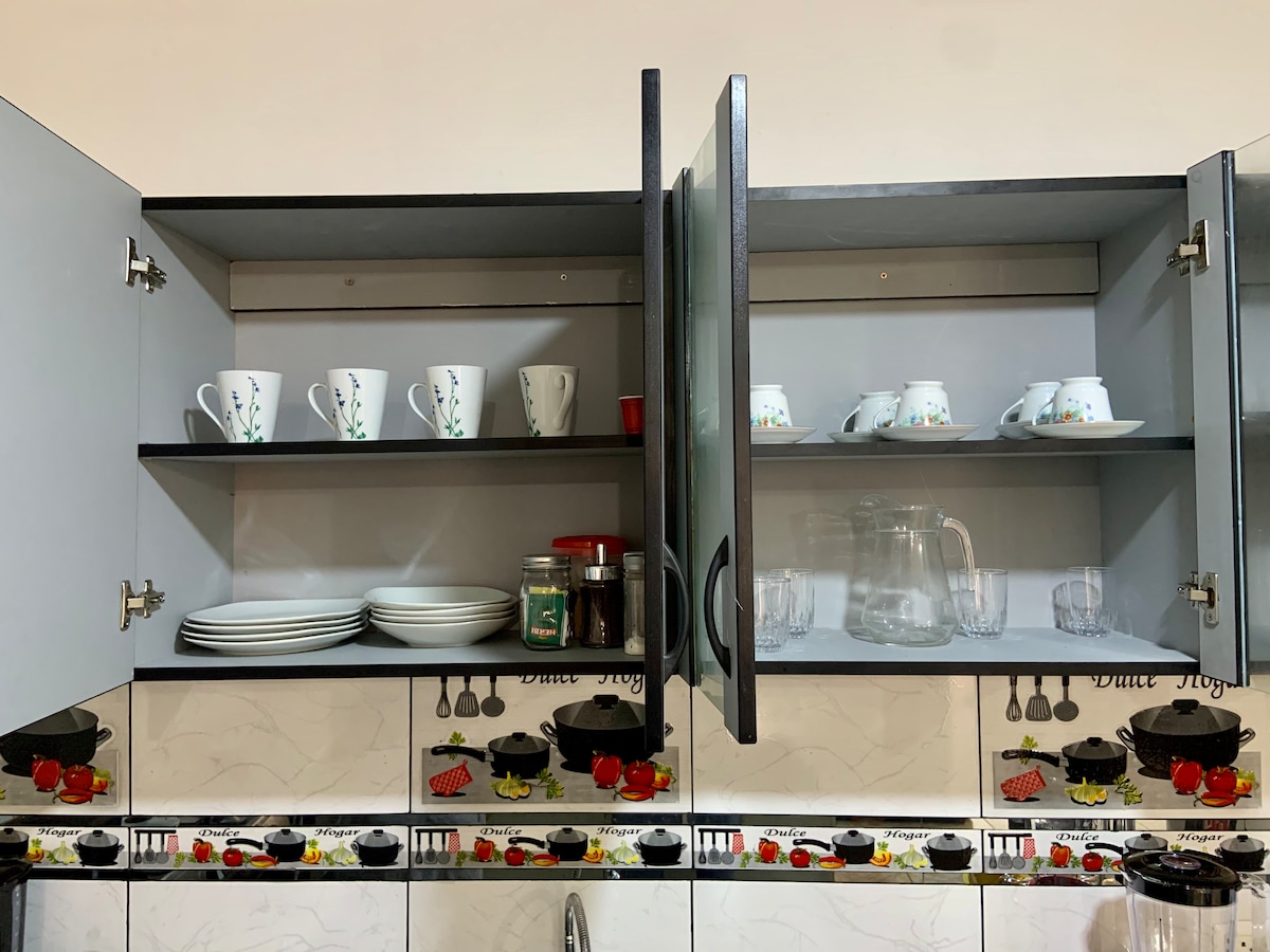 Upper kitchen cabinets are open, displaying a set of white dishes and cups with floral designs. Glass jars and a pitcher are visible, along with clear drinking glasses. The cabinets are complemented by a patterned backsplash featuring cooking imagery.