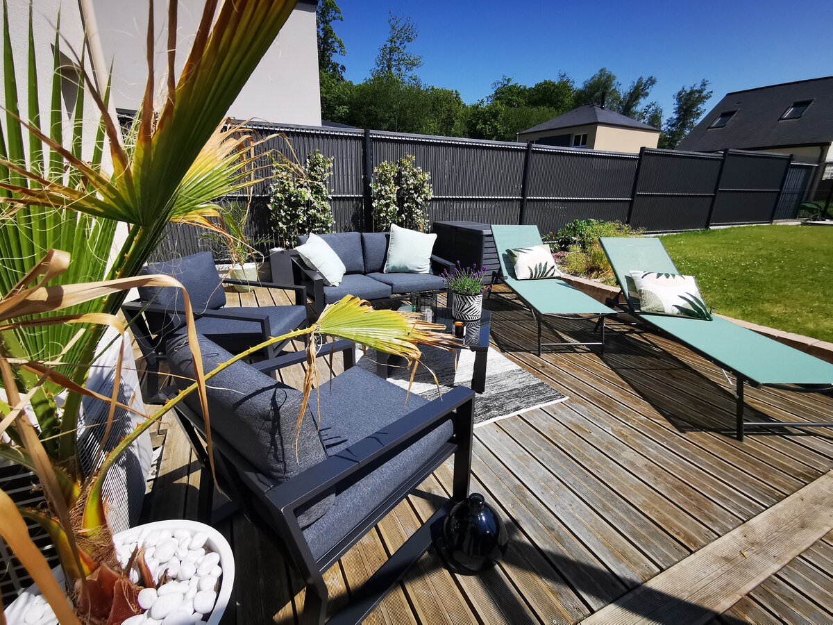 The outdoor terrace features comfortable seating arrangements, including a lounge set and two sun loungers. Lush greenery and decorative plants enhance the space. The wooden deck reflects the sunlight, creating a relaxing environment for guests to enjoy.