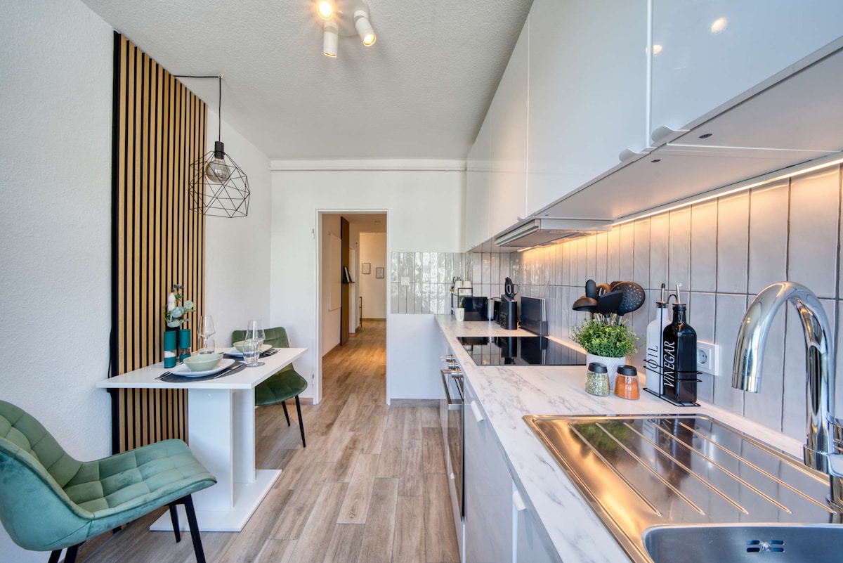 A modern kitchen space showcases sleek cabinetry and a marble countertop. A small dining table for two is set with tableware, accompanied by two green chairs. Soft lighting enhances the bright atmosphere, complementing the kitchen's full appliances and contemporary design.
