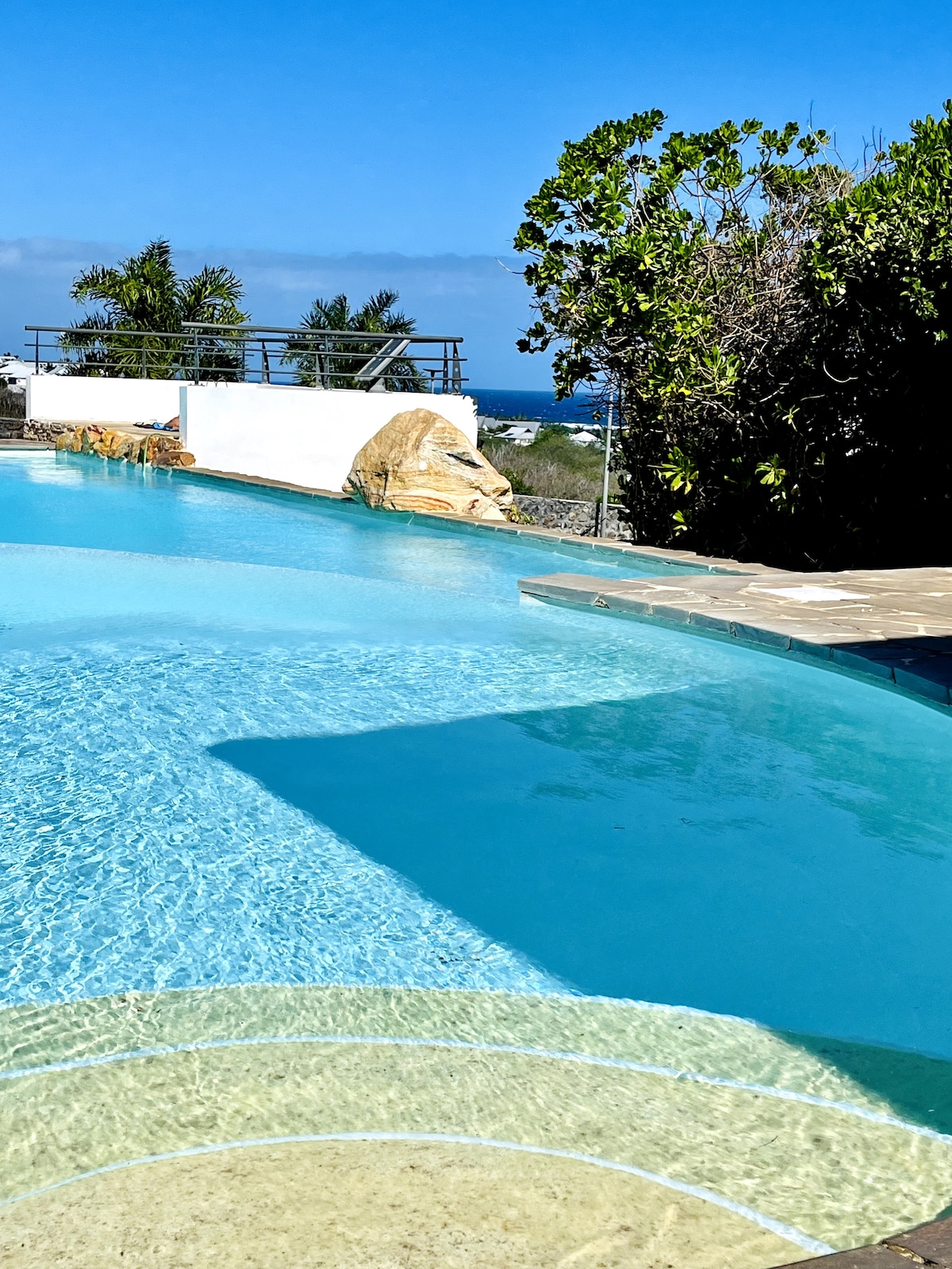 The image captures a serene swimming pool, with clear blue water reflecting the sky. Lush greenery borders the area, and the ocean horizon is visible in the background, enhancing the tranquil setting.