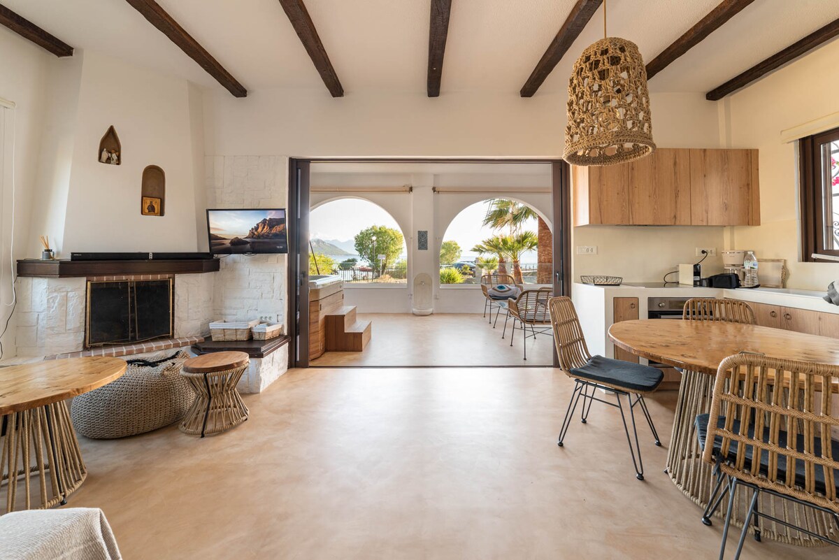 An open living area is defined by wooden beams and terracotta accents. Natural light floods in through large arches, highlighting a cozy seating arrangement and a fully equipped kitchen. Decorative elements include a woven chandelier and two round dining tables, creating a relaxed atmosphere.