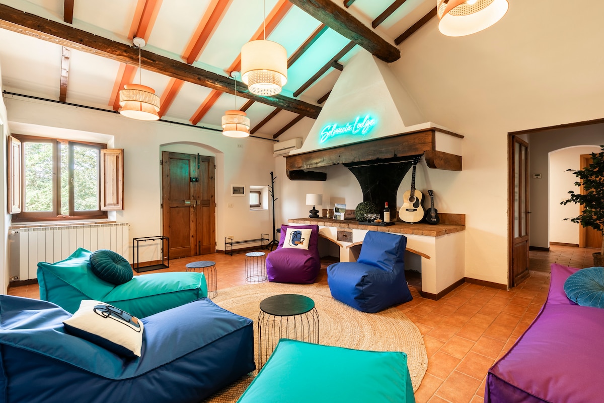 A shared living room features colorful bean bag chairs arranged around circular tables. A cozy area rug anchors the space, complemented by wooden beams and ceiling lights. Natural light fills the room through multiple windows, enhancing the inviting environment.