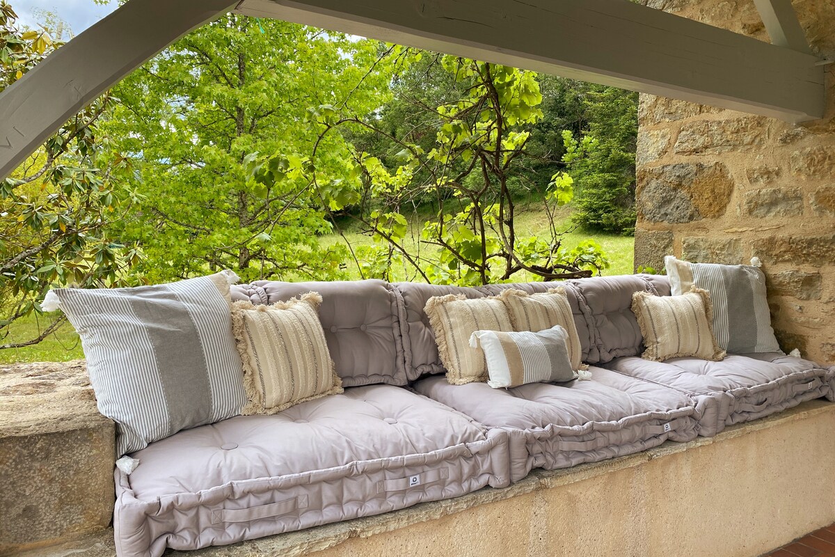 A comfortable seating area is positioned under a covered terrace. Soft cushions in light neutral tones are arranged along a stone bench, providing an inviting space to relax. Lush greenery is visible just beyond, enhancing the tranquil outdoor setting.