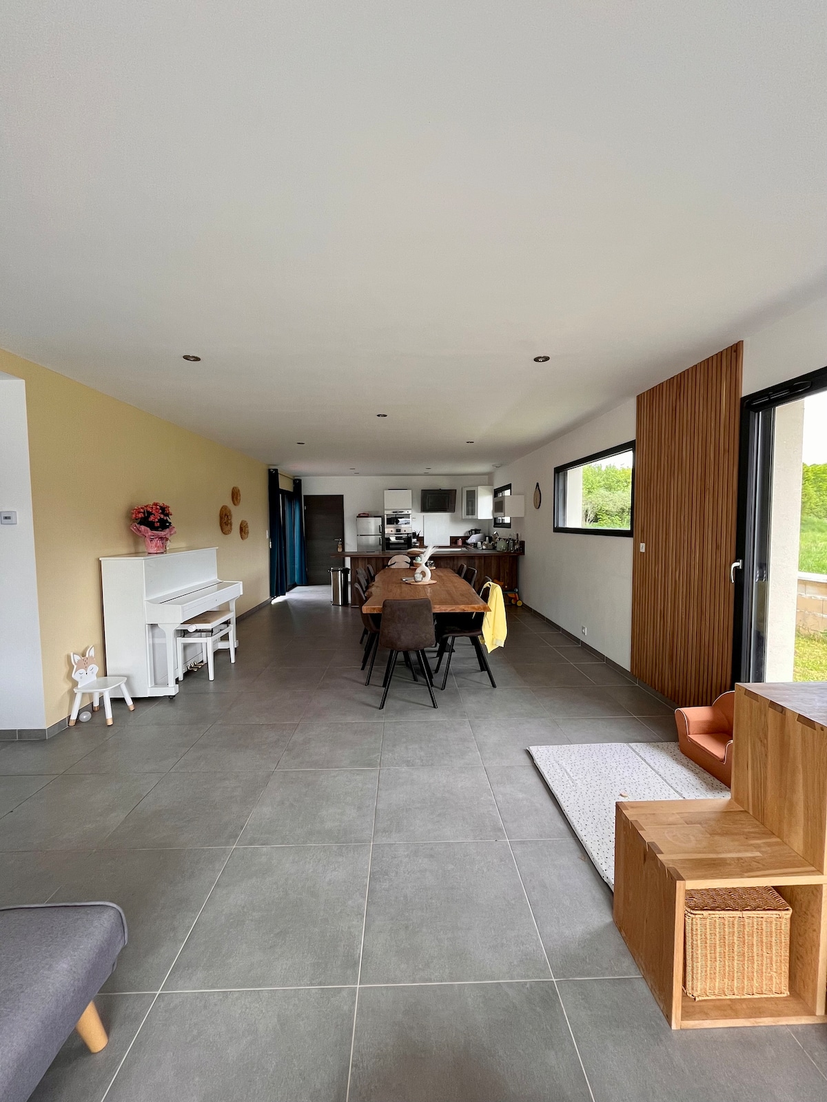 An open-plan living space is presented, featuring a dining area with a long table and several chairs. A kitchenette is visible in the background, with light entering through large sliding doors. Elements of warm wood and soft tones create a welcoming ambiance.