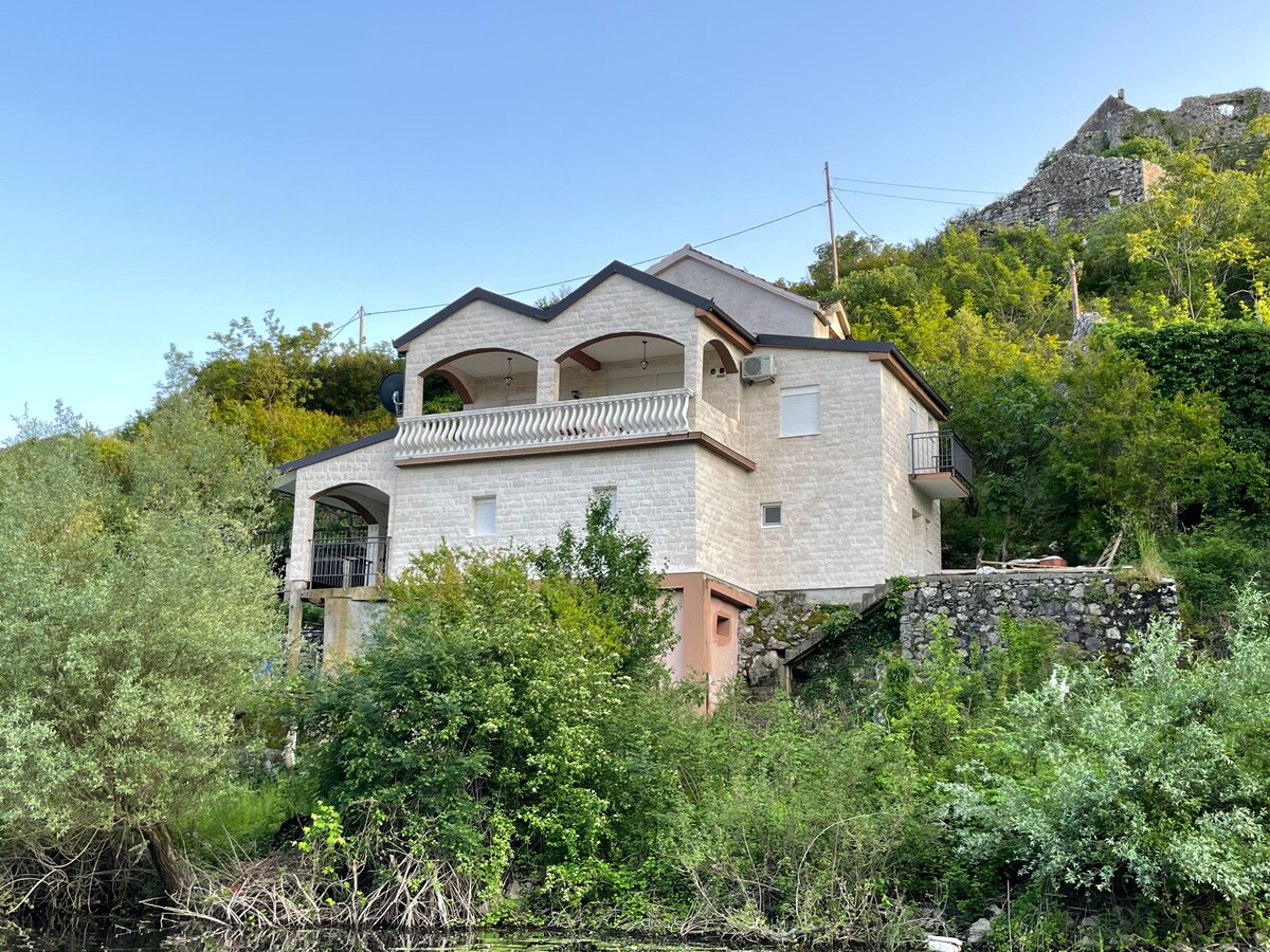 A two-story building is situated along the water's edge, featuring a light-colored stone facade. Multiple balconies with archways offer views of the surrounding greenery. Lush shrubs and trees are present in the foreground, contributing to a natural setting near the lake.