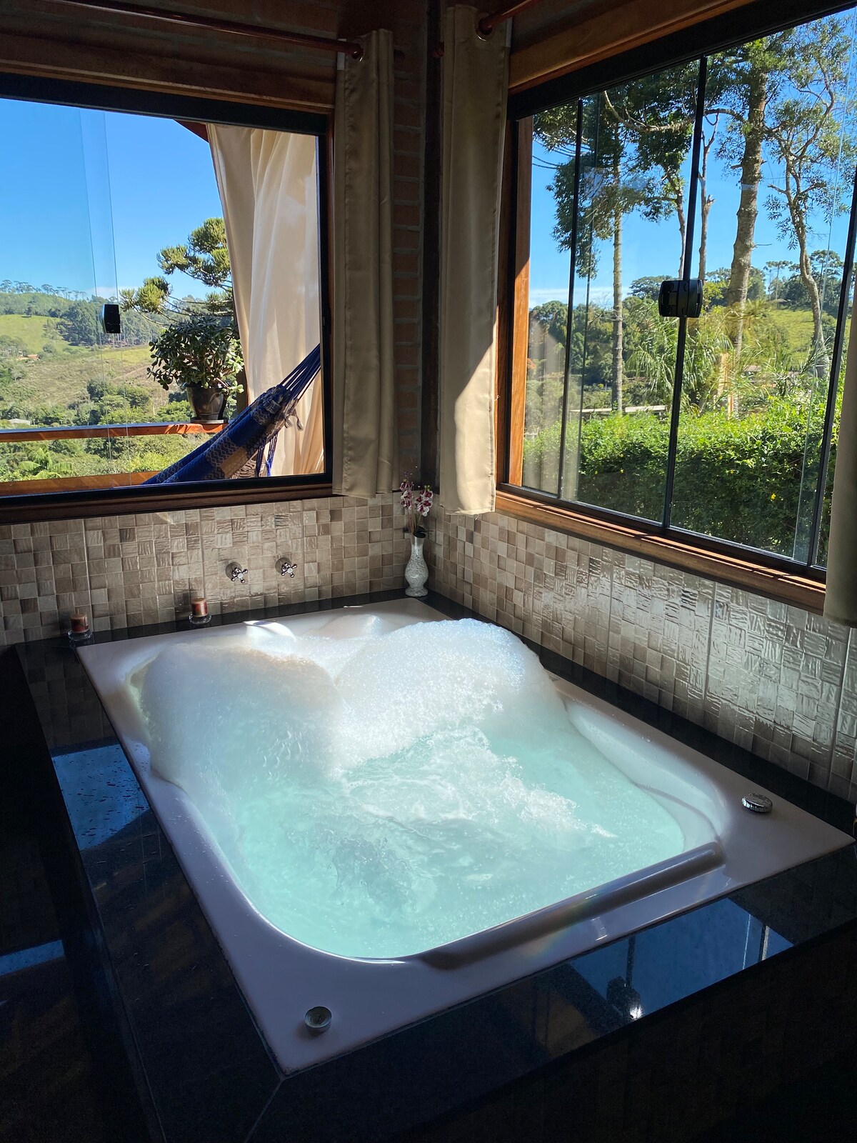 A spacious hydromassage bathtub is positioned beside large windows, providing views of the surrounding nature. The room features a tiled backsplash and allows natural light to enhance the tranquil atmosphere. Bubbles float on the surface of the water, inviting relaxation.
