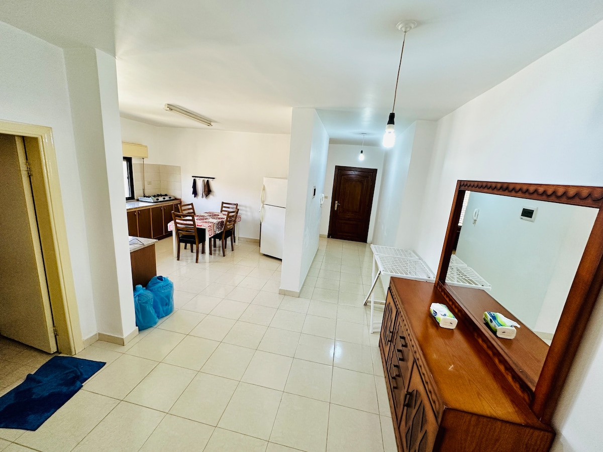 A bright and spacious living area is showcased, featuring a dining table surrounded by several chairs. The kitchen area is visible in the background, equipped with essential appliances. A wooden sideboard is positioned against the wall, complemented by a minimalistic decor on top.