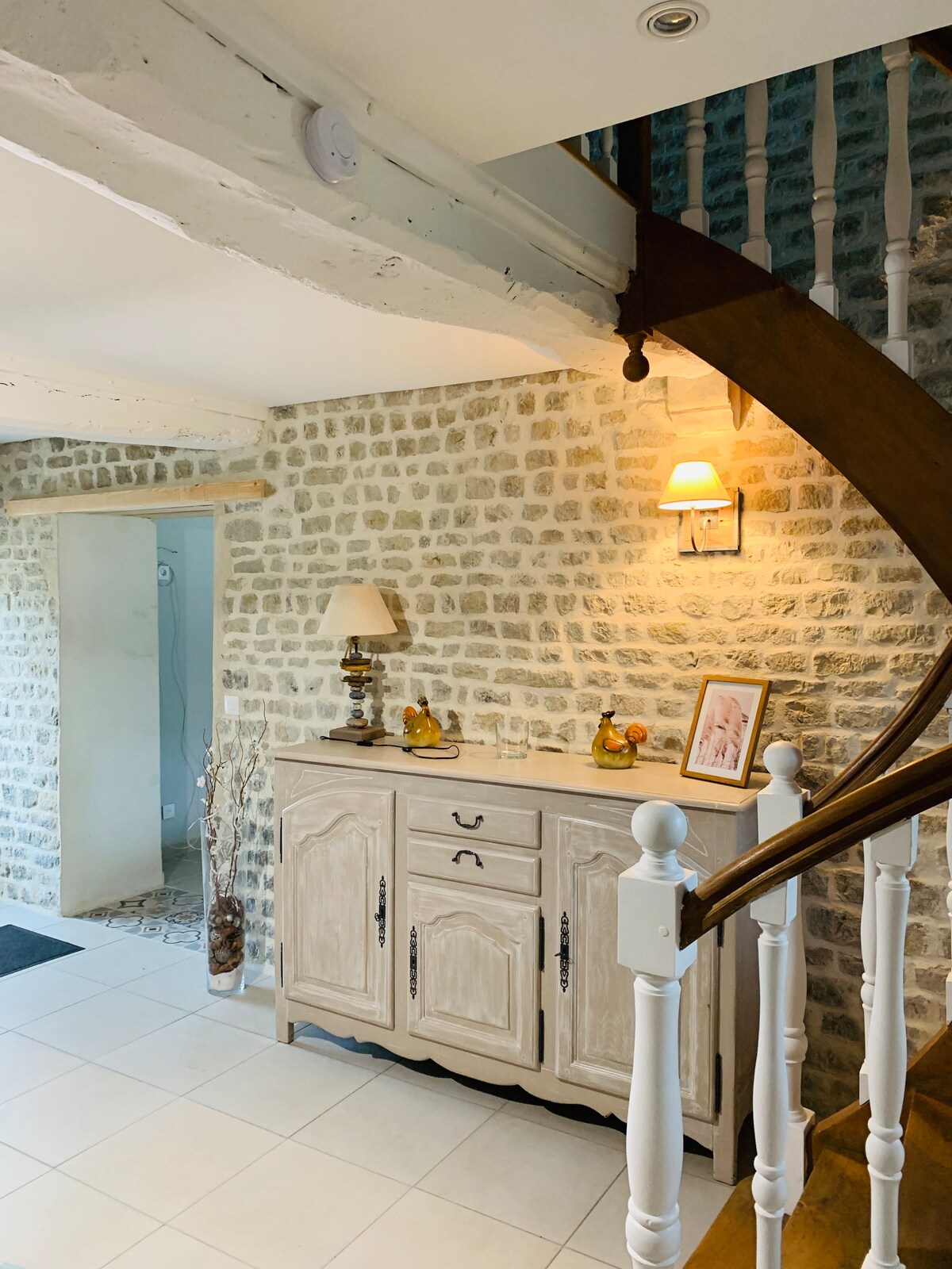 An entryway features a light stone wall and a wooden staircase leading to the upper level. A chest of drawers, styled in a soft hue, is positioned against the wall. A lamp provides soft illumination, with decorative accents displayed atop the chest.