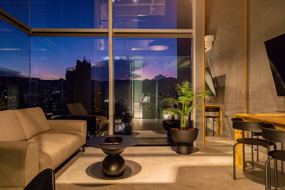 A contemporary living area features a comfortable sofa and a round coffee table, with large glass windows offering expansive views of the city skyline at dusk. Soft lighting creates a calming ambiance, complemented by a green plant beside the window.