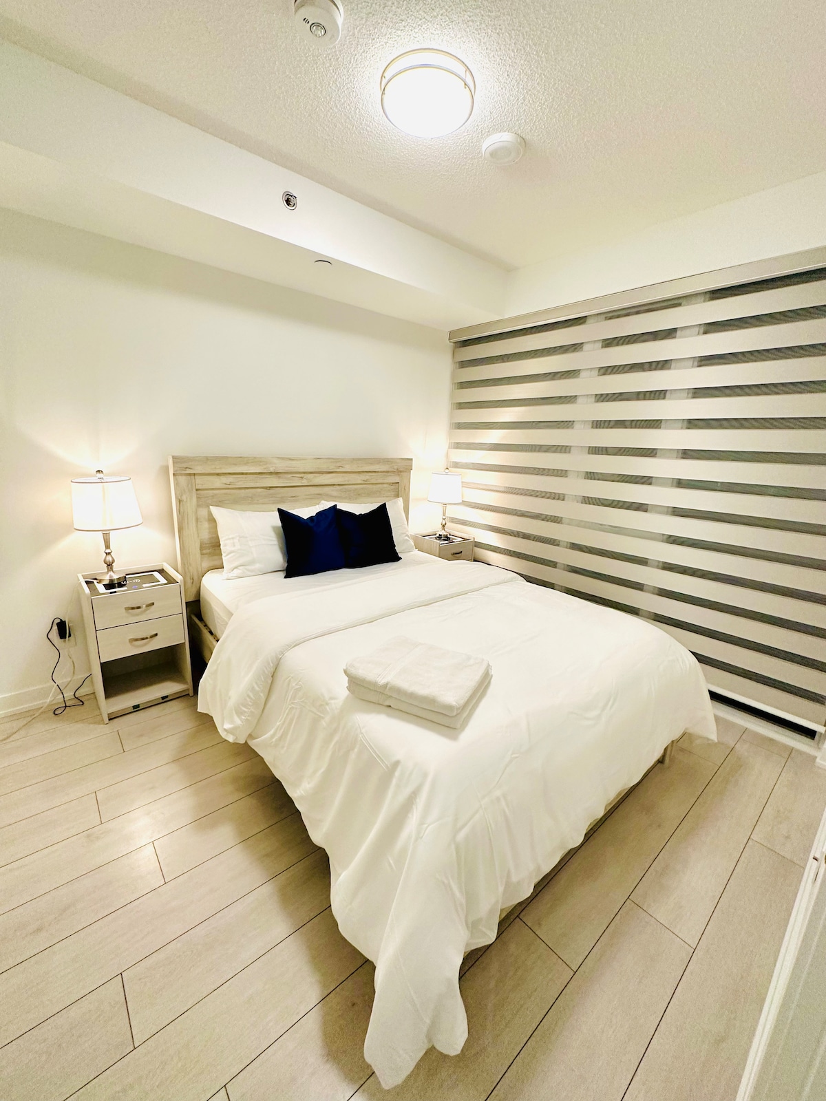 The bedroom features a spacious bed with a white duvet and a decorative blue pillow. Two bedside lamps provide soft lighting beside the bed. A nightstand is positioned on one side, while striped window treatments allow natural light to enter while ensuring privacy.