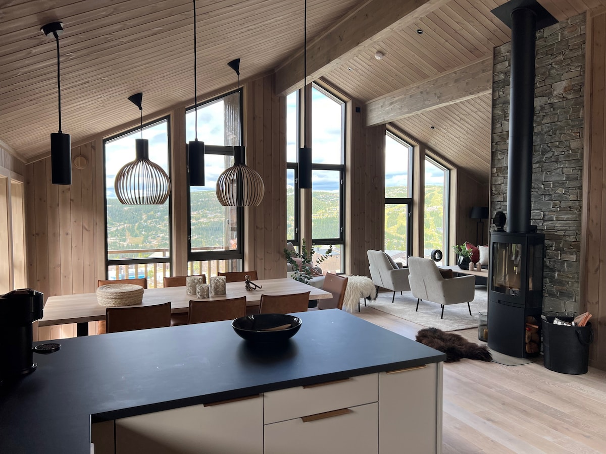 An open living space is highlighted by large windows offering expansive views of the surrounding landscape. A black kitchen island contrasts with light wood walls, while a dining table is set for several guests. A cozy seating area includes modern armchairs and a wood-burning stove.