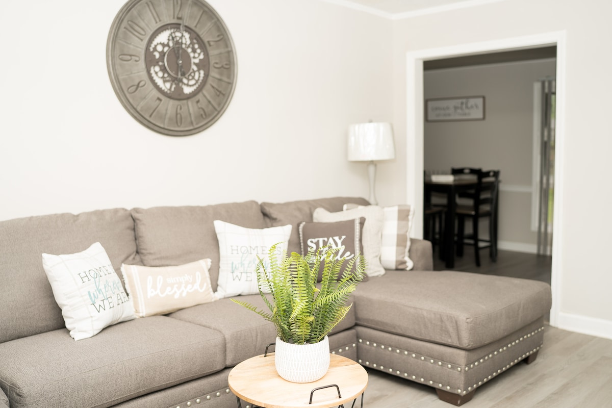 A comfortable seating area features a gray sectional sofa adorned with decorative pillows displaying various texts. A round wooden table with a plant sits in front, while a wall clock is mounted above the sofa. Subtle lighting enhances the inviting space.