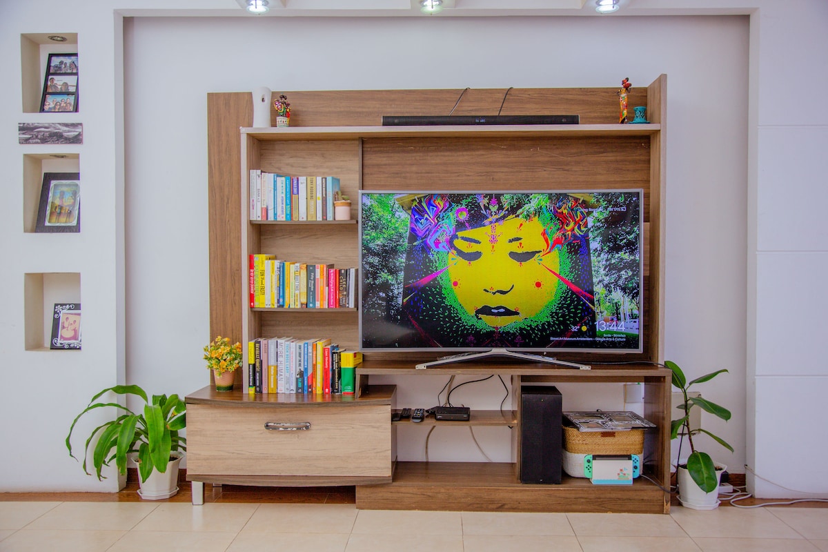 A modern entertainment unit features a large television displaying colorful art. Surrounding the TV, shelves are lined with books, while decorative plants add greenery. A low cabinet sits beneath, providing additional storage space.