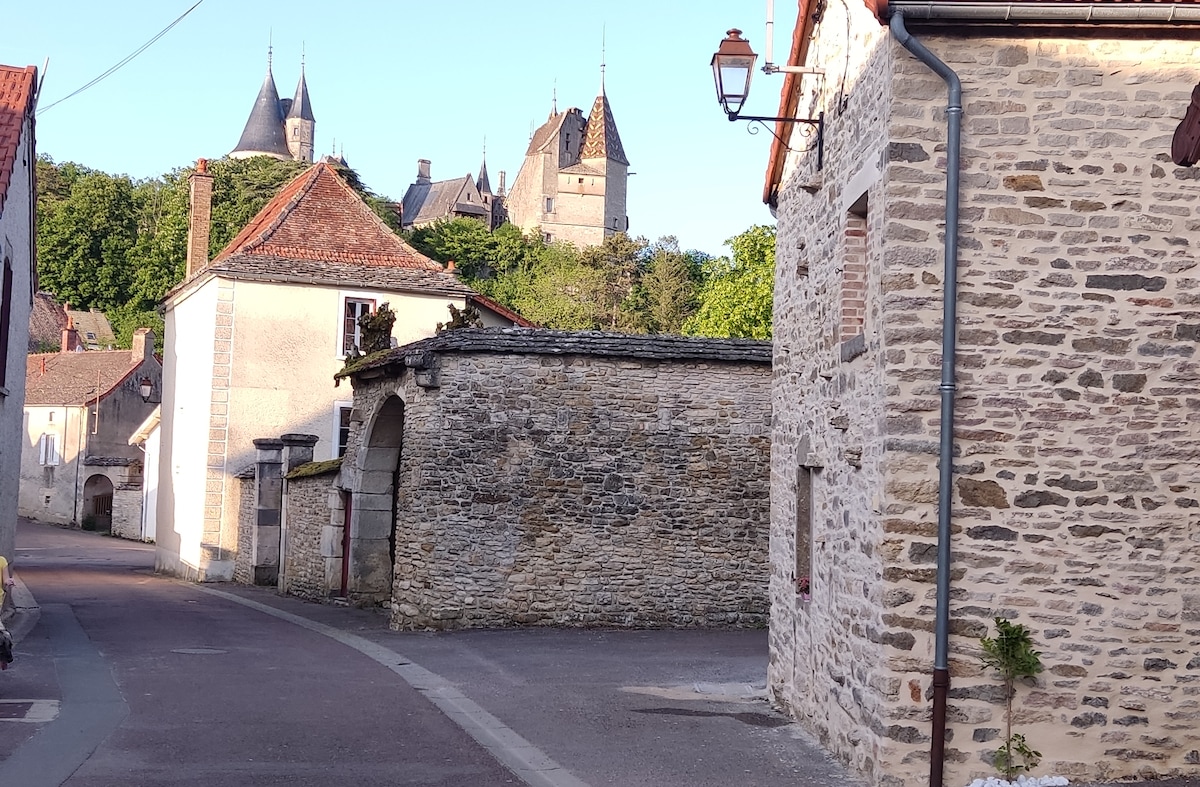A peaceful village street is lined with stone buildings that reflect traditional architectural styles. In the background, a historic castle with turrets and a lush green hillside creates a scenic backdrop, enhancing the charming ambiance of the area.