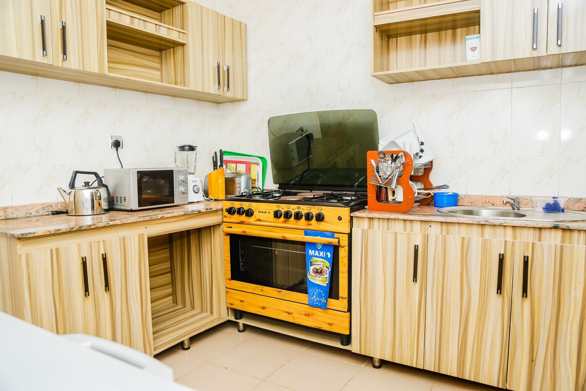 A well-equipped kitchen features wooden cabinetry and ample countertop space. An oven and stovetop are positioned on one side, with a microwave and kettle nearby. Various kitchen accessories, including a mixer, are organized on the counter, offering convenience for family meal preparation.