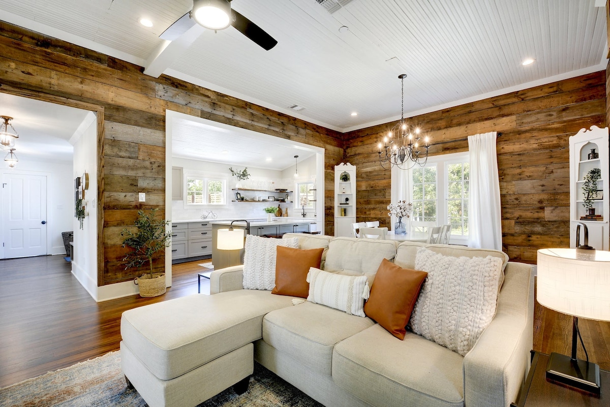An open living area features a light-colored sectional sofa adorned with orange and cream cushions. Warm wood paneling contrasts with the bright wall, and a dining area is visible in the background. Natural light floods the space from multiple windows.