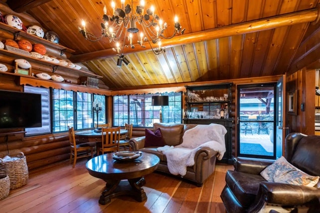 A warm living area features wooden walls and ceiling beams, with a large circular table surrounded by dining chairs. A plush sofa and armchair are positioned beside a rustic bookshelf. Natural light streams in through expansive windows, enhancing the cozy atmosphere.