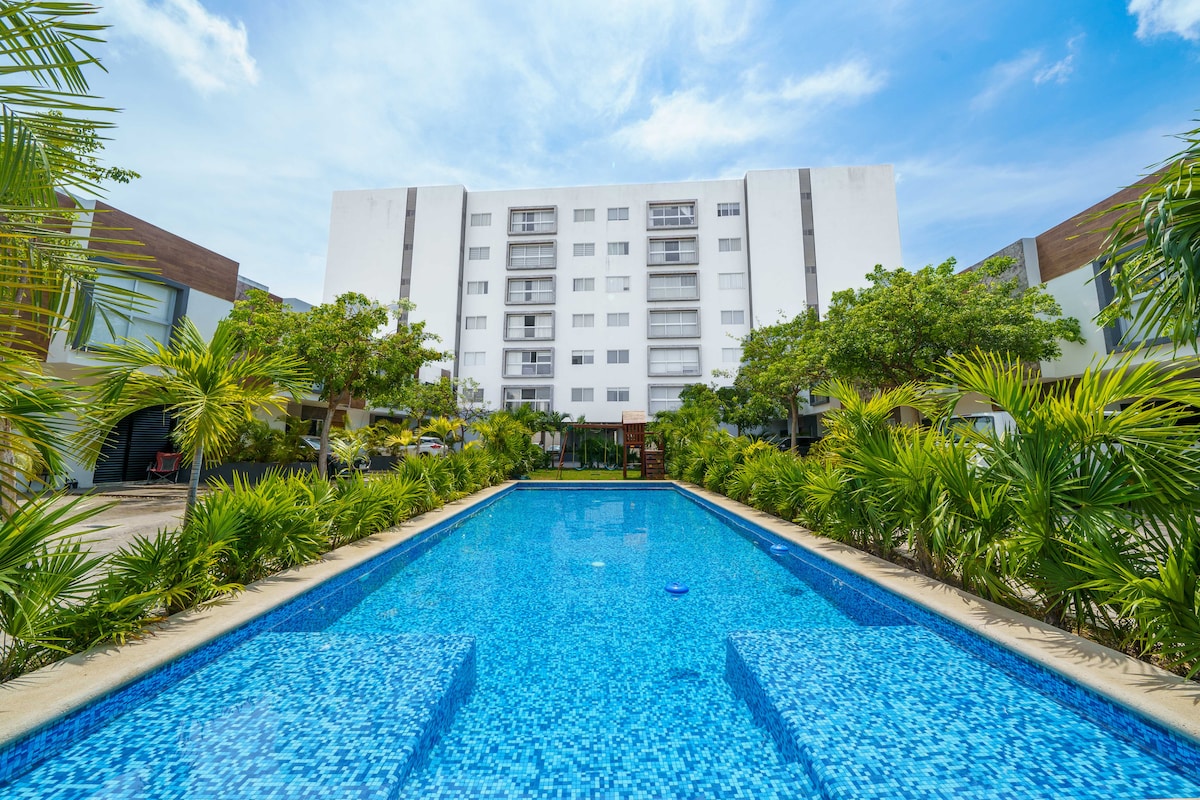 A serene swimming pool is surrounded by lush greenery, with a modern apartment building in the background. The pool features bright blue tiles and is designed for relaxation under clear skies.