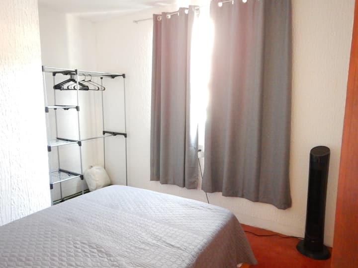 Izba na prízemí s nábytkom na oblečenie, stolom, háčikmi, zatemňovacou závesou, ventilátorom, odpadkovým košom a ďalšou prikrývkou v priestore na vešanie oblečenia 