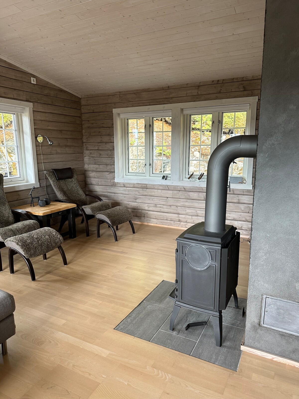 A cozy living area features a wood-burning stove, enhancing warmth during cooler months. Comfortable seating is provided by two armchairs and a small table, with natural light entering through multiple windows framed by wooden walls.
