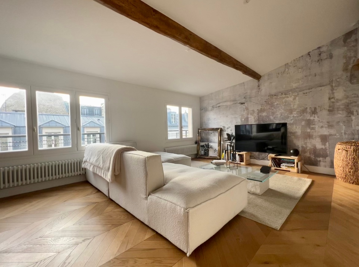 The bright living room features a spacious corner sofa in soft fabric, complemented by a large television. Natural light fills the space, enhanced by multiple windows overlooking the skyline. A stylish coffee table rests on a light-colored rug over wooden parquet flooring.