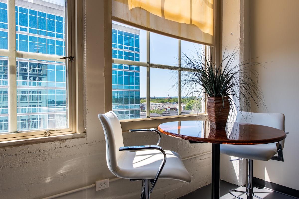 A small dining area is illuminated by natural light, featuring a wooden table surrounded by modern white chairs. A potted plant adds a touch of greenery, while large windows showcase a view of the city skyline and distant landscape.