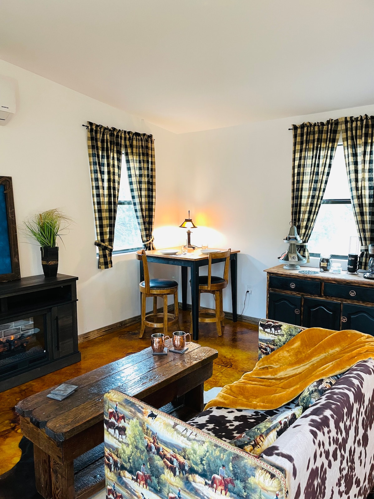 A cozy seating area is highlighted by a cowhide-patterned couch and rustic wooden furniture. Natural light filters through the matching checkered curtains at the windows. A small table with two stools and a decorative lamp create a warm space, complemented by a compact electric fireplace.