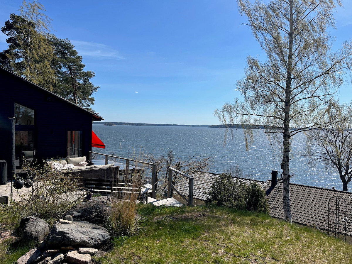 A spacious outdoor area features a view of the water, framed by greenery and a birch tree. The patio is equipped with seating and a sun umbrella, inviting relaxation. The surrounding landscape includes natural rock formations and well-maintained grass.