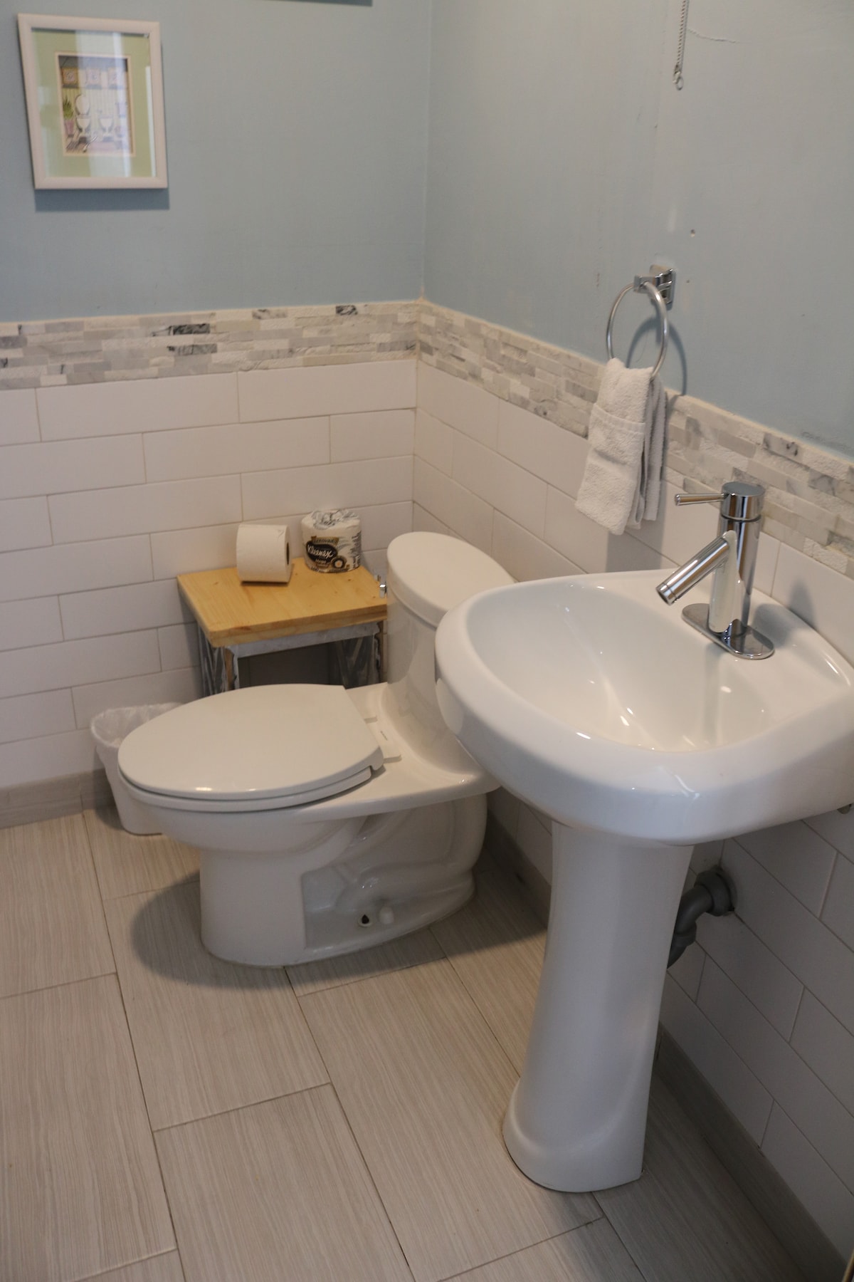 A compact bathroom is featured with modern fixtures. A white pedestal sink is situated next to a toilet. A small wooden table displays toiletries, while patterned tiles add subtle texture to the walls. Light-colored tile flooring enhances the space's brightness.