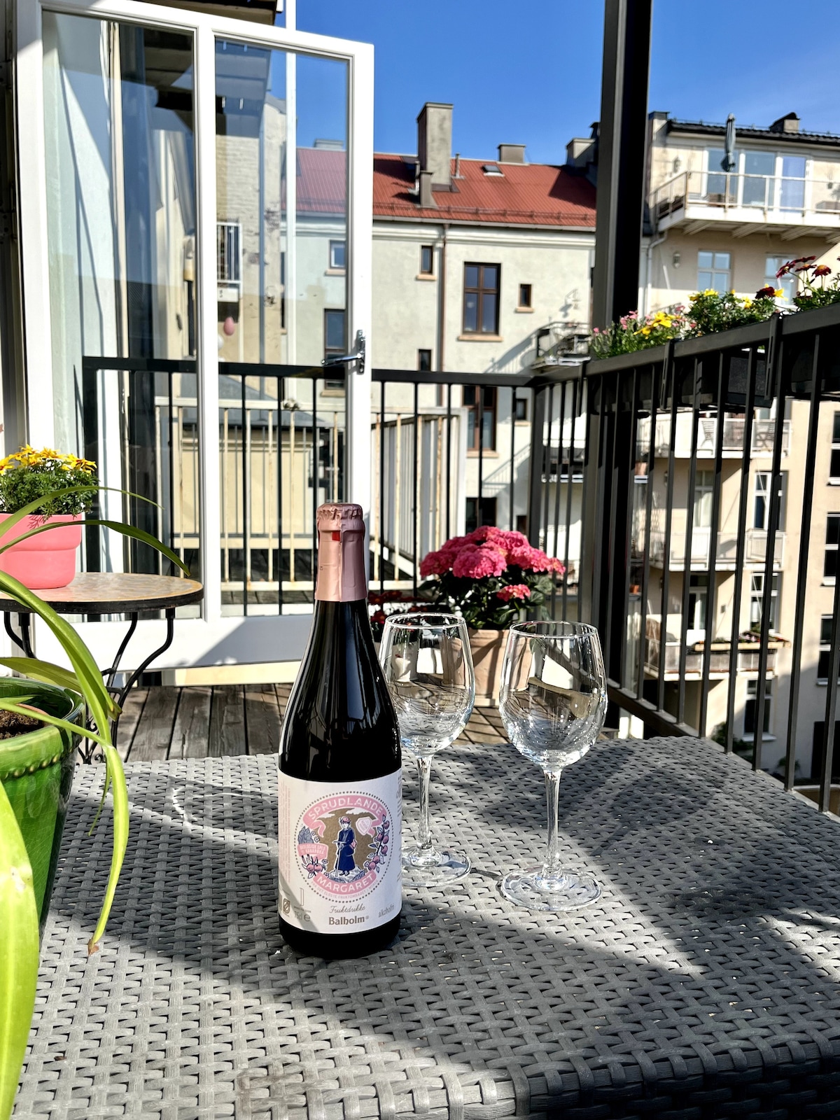 A balcony is presented with a bottle of wine and two glasses, resting on a wicker table. Colorful flowers are seen in pots, and the backdrop features a view of surrounding buildings under a clear blue sky.
