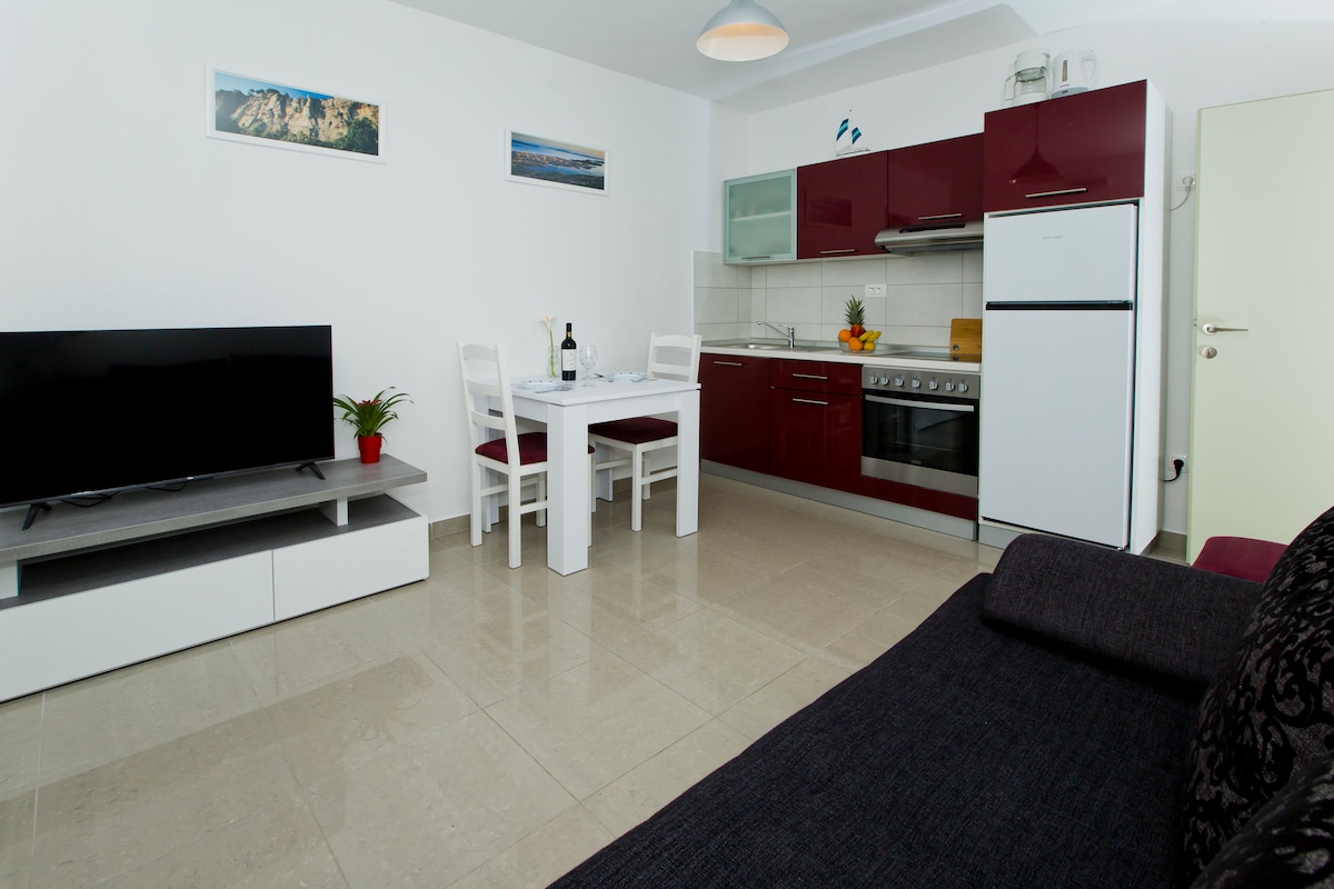 A modern living space is visible, featuring a sleek kitchen with red cabinets and silver appliances. A dining table is arranged for four, complemented by white chairs. A television sits on a low white unit, while a dark sofa provides seating, all on a polished tile floor.