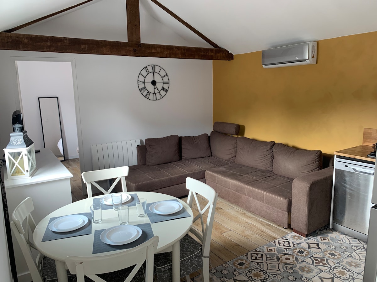 A spacious living area features a brown sectional sofa, accommodating multiple guests. A round dining table is set with plates and glasses, complemented by white chairs. Exposed wooden beams add character to the high ceiling, while a subtle decorative clock enhances the ambiance.