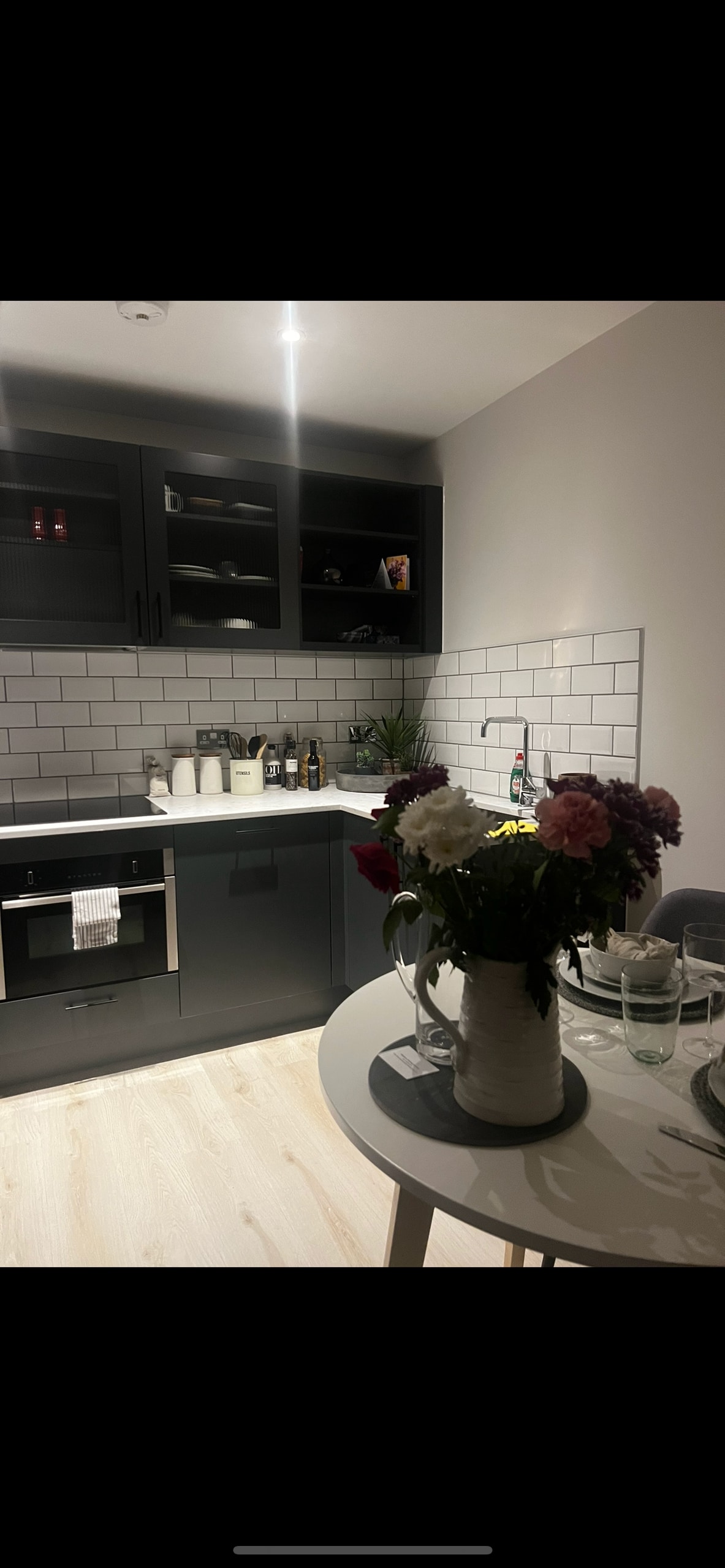A modern kitchen is equipped with sleek black cabinetry and white tile backsplash. A round dining table is set nearby, featuring a vase of vibrant flowers and glassware. Warm overhead lighting enhances the clean, inviting atmosphere.