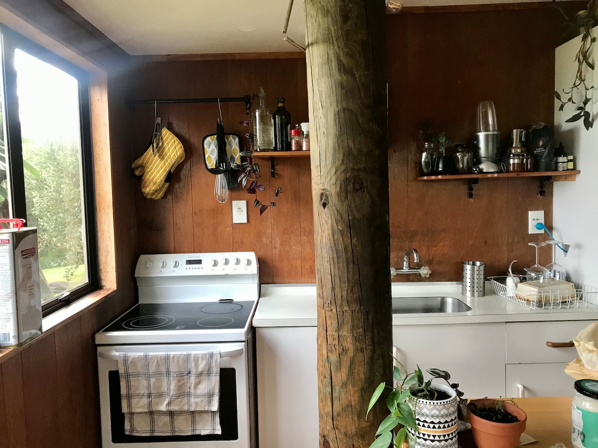 The kitchen features wooden paneling and a large window providing natural light. A stove and sink are visible, along with shelves displaying kitchenware and utensils. Potholders hang from the wall, and a wooden post supports the ceiling, adding to the rustic feel.