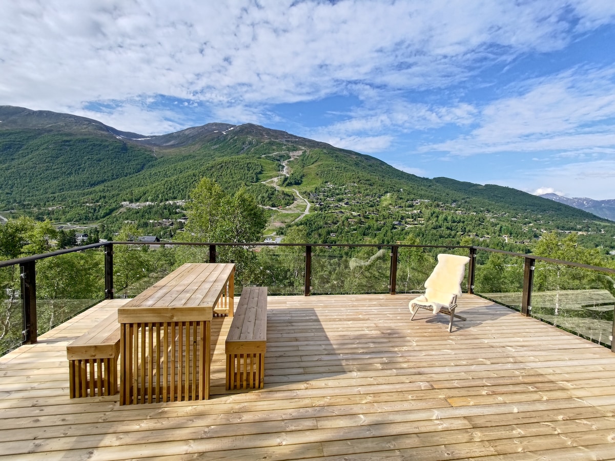 An outdoor terrace features a large wooden dining table surrounded by benches. A single lounge chair is placed nearby. The space offers expansive views of green mountains and ski slopes under a clear blue sky, creating a peaceful setting for relaxation.