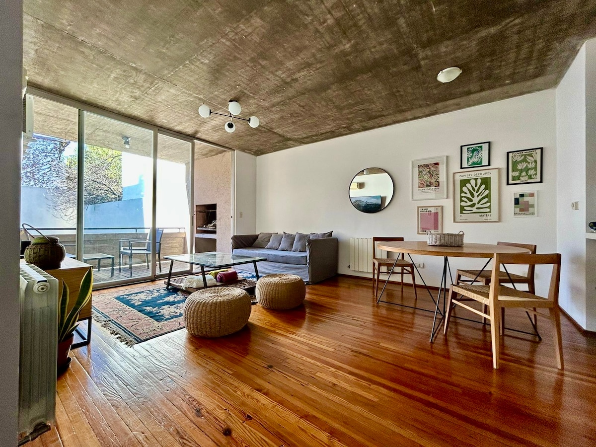 A modern living area is presented with a comfortable gray sofa facing large glass doors that open to a balcony. A round mirror and various framed artworks decorate the walls. A dining table with four chairs complements two woven ottomans on the wooden floor.