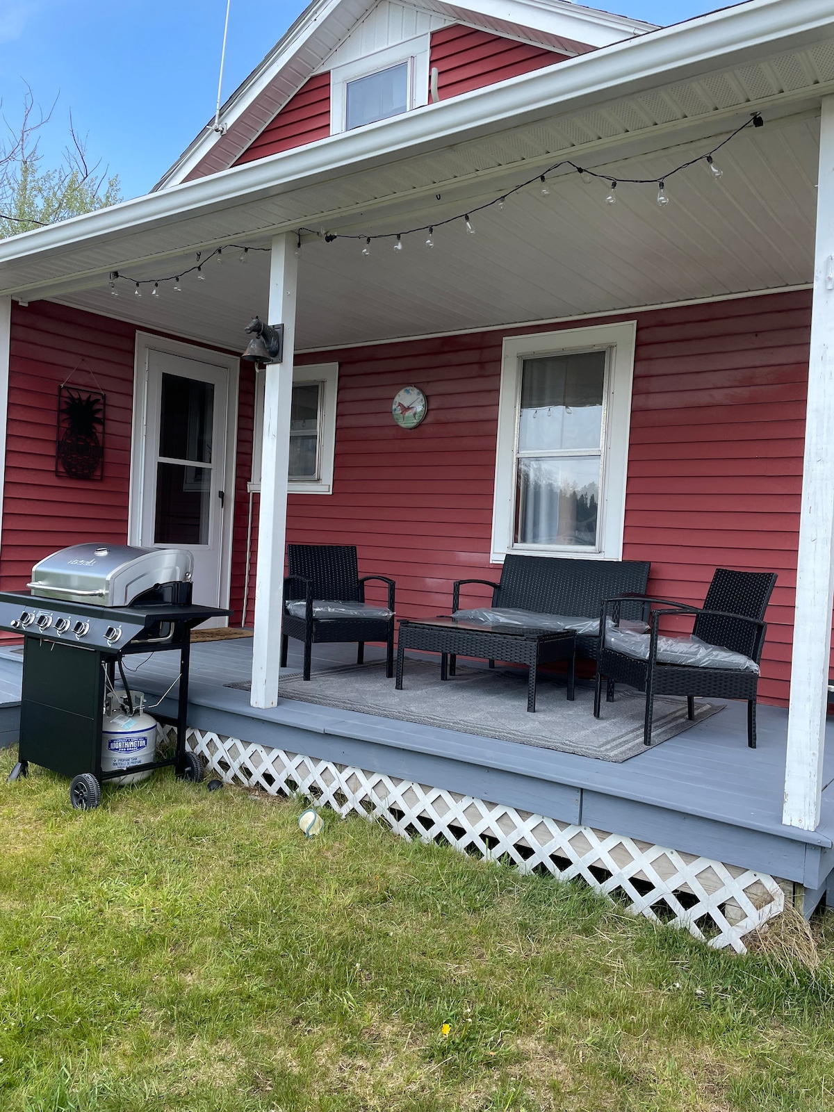 A covered porch features a gas grill beside a small dining set, accompanied by four black wicker chairs on a gray platform. String lights are positioned along the roof, creating a comfortable outdoor space against the backdrop of a red farmhouse.